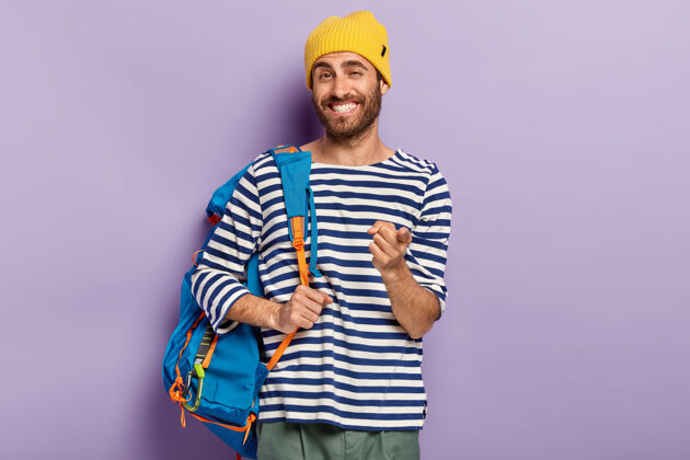 一个快乐微笑的男人旅行者的照片用食指指着你 背着背包 戴着黄色帽子和毛衣 表达选择 接你 孤立在紫色的墙上年轻徒步旅行者白种人