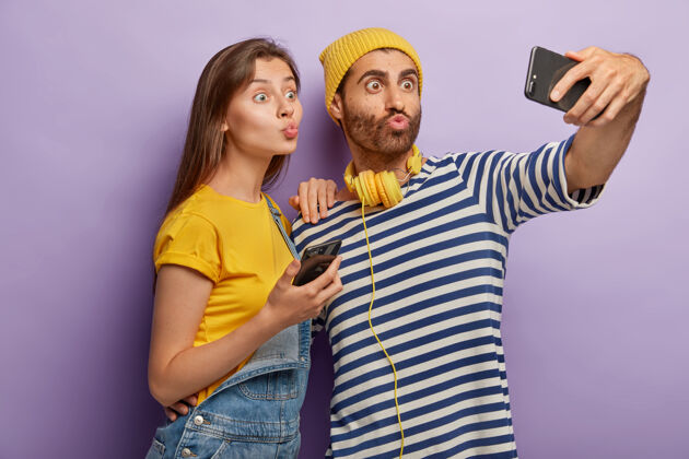 紫色搞笑的年轻男女做鬼脸 嘴唇圆润 用现代手机的前置摄像头拍照 自拍紫罗兰工作服室内