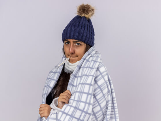 帽子年轻生病的女孩看着镜头戴着冬天的帽子用格子布裹着围巾把体温计放在嘴里隔离在白色的相机白色嘴巴