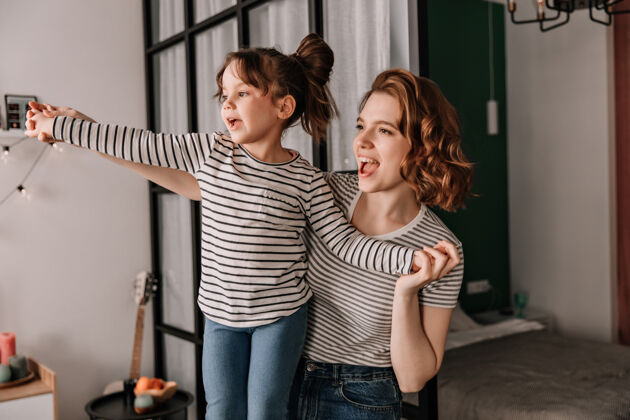 小积极的女人和她穿着条纹t恤的女儿在公寓里欢笑跳舞护理欢呼漂亮