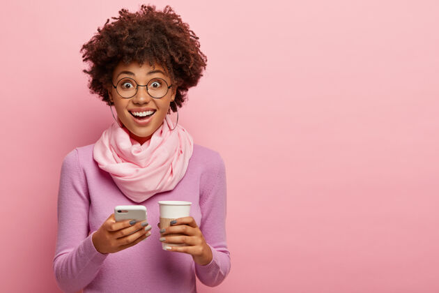服装年轻漂亮的黑皮肤女人 非洲式发型 手持手机和一次性咖啡杯 穿着紫色马球衫和围巾 在粉红色的墙上摆姿势通讯 生活方式 科技高兴一个一次性