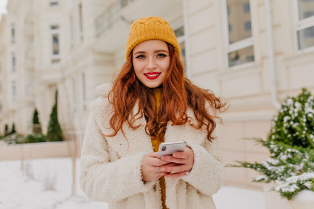 手机穿着针织帽子拿着电话的迷人白种女人穿着白色外套的姜汁女孩的户外照片冬天女孩配件