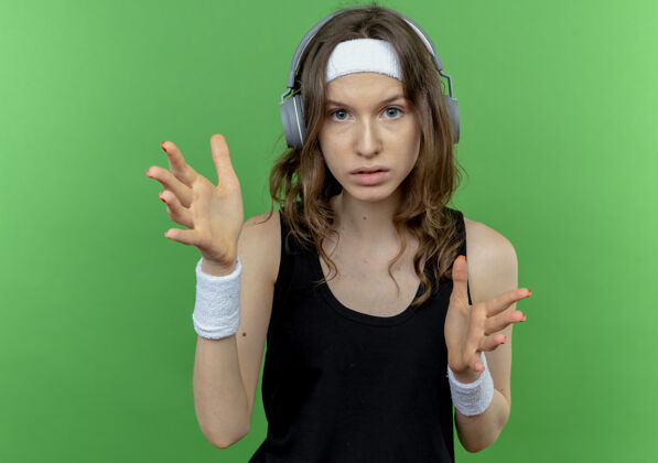 女孩身穿黑色运动服 头戴耳机 表情严肃 双手站在绿色墙壁上的年轻健身女孩健身手脸