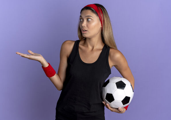 站身穿黑色运动服 头戴红色头巾 手举足球的年轻健身女孩站在蓝色的墙上忧心忡忡手臂球担心