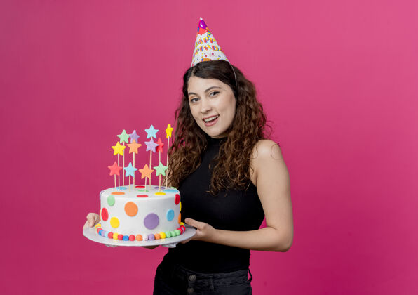 帽子年轻漂亮的女人 卷发 戴着节日帽 手里拿着生日蛋糕 站在粉色的墙上 快乐而积极的生日派对理念年轻积极女人
