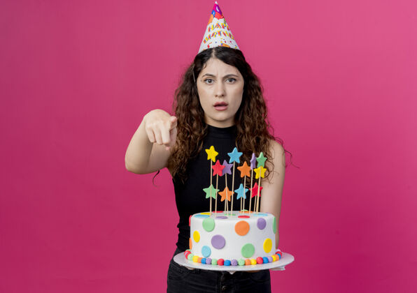 卷发一个年轻漂亮的女人 卷发 戴着节日帽 拿着生日蛋糕 用手指着 站在粉色的墙上 不高兴地庆祝生日站着不高兴蛋糕