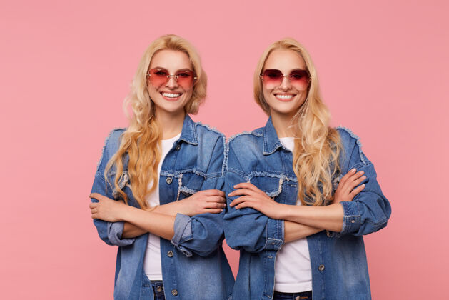 女士两位戴着太阳镜 穿着牛仔裤外套的白头美女双手交叉放在胸前 站在粉色的背景下 面带微笑 愉快地看着镜头姿势白皙姐妹