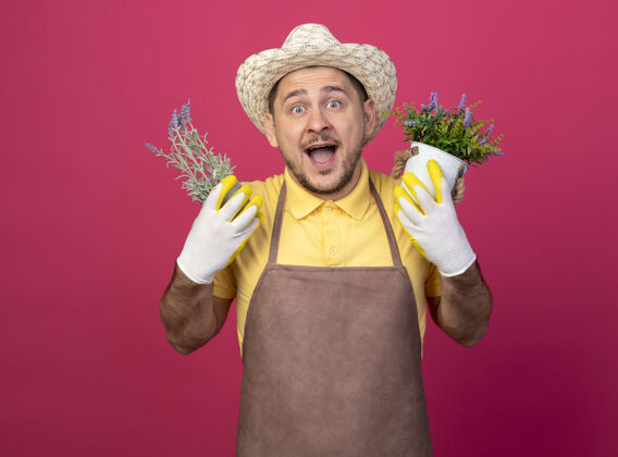站着年轻的园丁 穿着连体衣 戴着帽子 戴着工作手套 手里拿着盆栽植物 快乐快乐穿手套