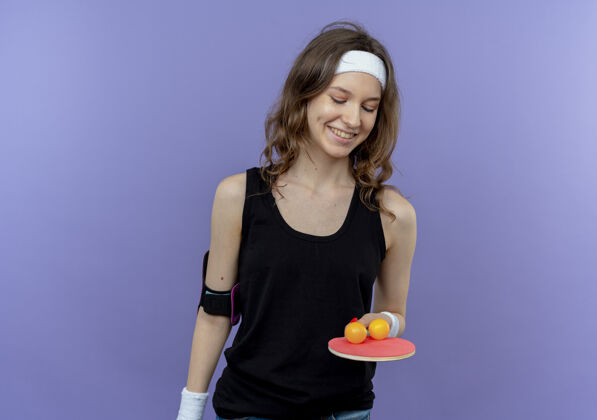 球拍身穿黑色运动服的年轻健身女孩 头上戴着头巾 手里拿着球拍和乒乓球 站在蓝色的墙上 快乐而积极地微笑着女孩运动装健身