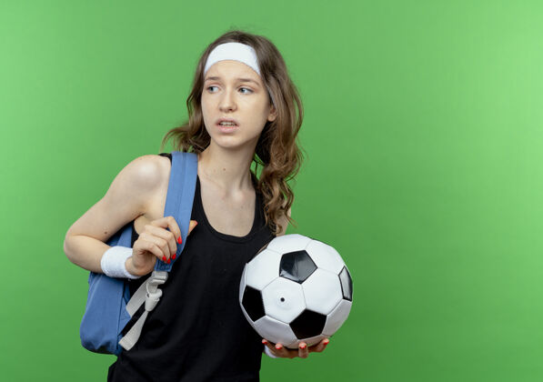 抱着穿着黑色运动服 背着背包 头戴足球带的年轻健身女孩站在绿色的墙上 茫然地看着一边健身背包女孩