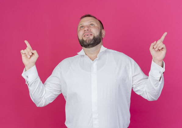 站着一个留着胡子的男人穿着白衬衫 食指朝上 站在粉色的墙上 开心而积极地微笑着手指目录穿