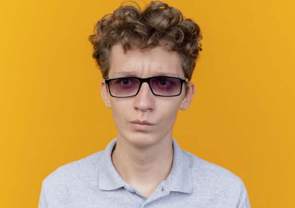 年轻一个戴着黑眼镜 穿着灰色马球衫的年轻人站在橘色的墙上 眉头紧锁地看着一边一边看穿