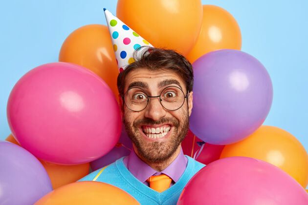 完美帅哥快乐的头像被派对气球包围着摆姿势帽子眼镜男人