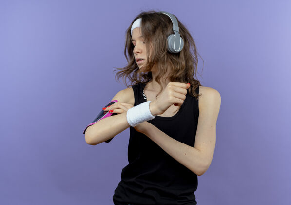 健身身穿黑色运动服的年轻健身女孩戴着头带 戴着耳机 手在蓝色的墙上固定着智能手机臂带头带耳机手