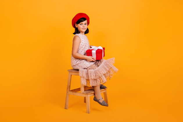 贝雷帽穿着裙子拿着生日礼物的未成年女孩带着礼物的孩子坐在黄色墙上的椅子上孩子表情乐趣