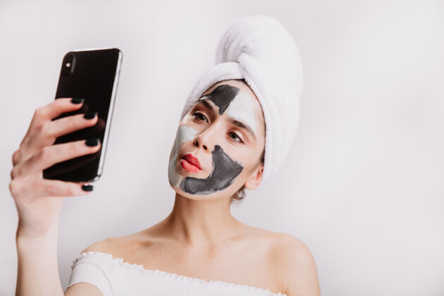 护肤一个滑稽的女人 戴着粘土面膜做面部护理 头上戴着毛巾 在白墙上自拍完美黏土光泽