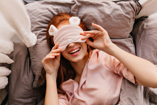 周末浪漫的女孩躺在床上 带着充满灵感的微笑幽默的姜女的头顶镜头睡眠面具红发年轻女士