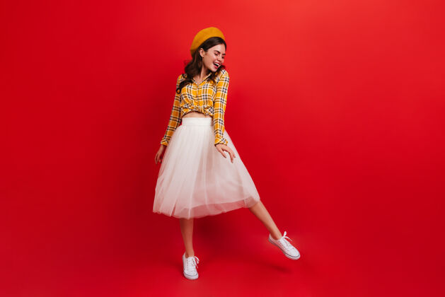 全长戴着时髦的橙色帽子 穿着鲜艳上衣的年轻女子正在红墙上跳舞穿着白色裙子和运动鞋的女孩微笑着黄色衬衫帽子肖像