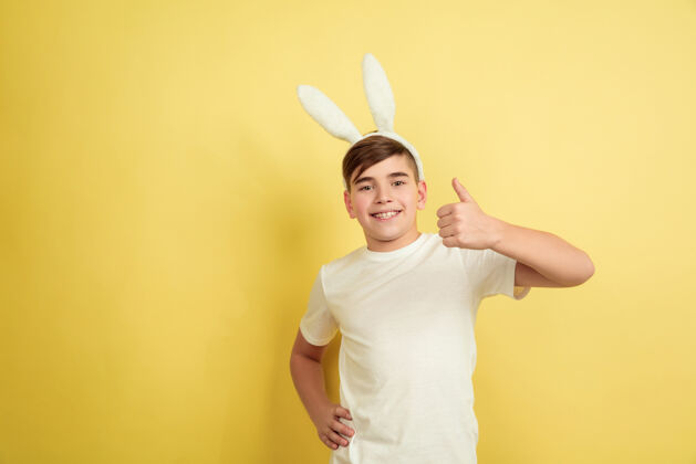 面部竖起大拇指白人男孩在黄色工作室背景上扮演复活节兔子复活节快乐乐趣复制空间姜