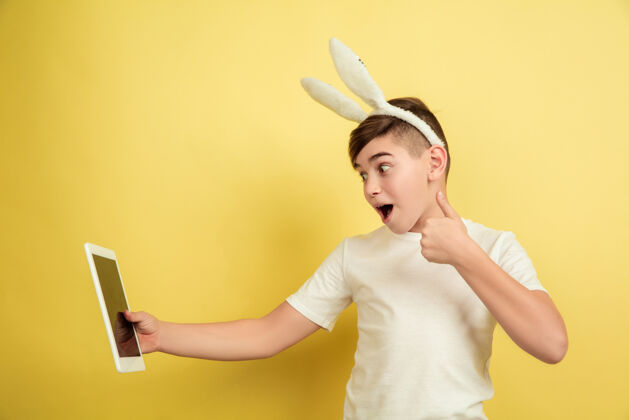 男性使用平板电脑 竖起大拇指白人男孩作为复活节兔子在黄色工作室背景复活节快乐问候五颜六色姜表情