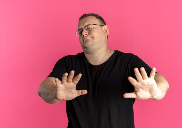 靠近一个戴眼镜的超重男人穿着黑色t恤 站在粉色的墙上停止唱歌 伸出手告诉大家不要靠近歌手穿做