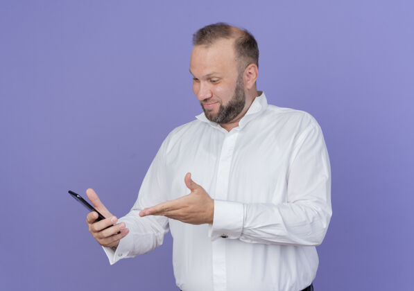 手机一个留着胡子的男人穿着白衬衫看着他的智能手机屏幕 手臂微笑着站在蓝色的墙上穿介绍看