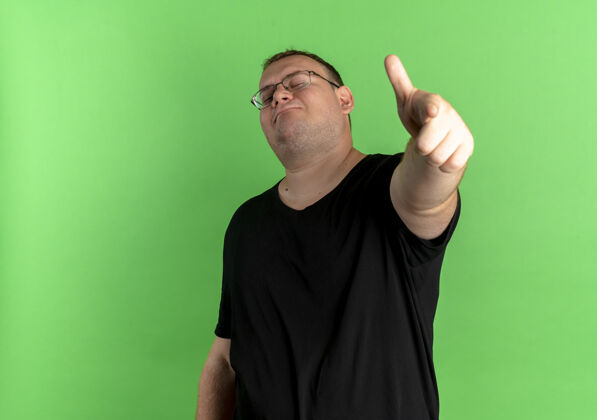 穿戴眼镜的超重男子身穿黑色t恤 看上去自信满满 食指站在绿色的墙上指眼镜超重