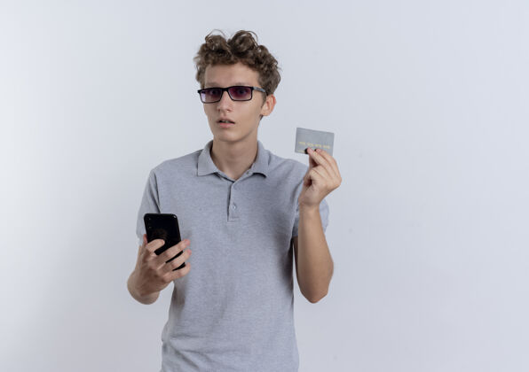 显示身穿灰色马球衫的年轻人手持智能手机 站在白墙上 显示信用卡被搞糊涂了信用举行移动