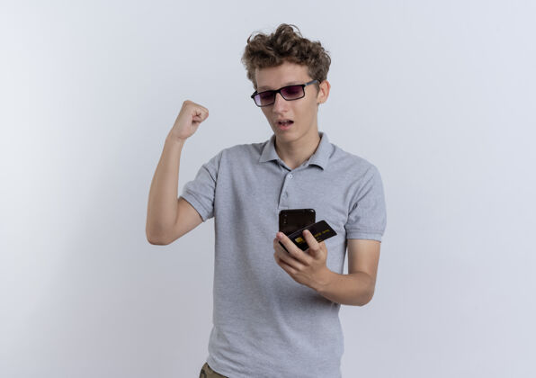 男人一个穿着灰色马球衫的年轻人站在白色的墙上看着他的智能手机屏幕 手里握着信用卡 握紧拳头 高兴而兴奋拳头移动信用