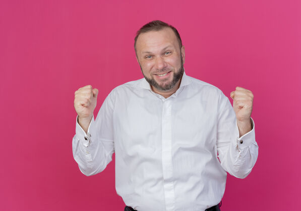 男人一个留着胡子的男人穿着白衬衫 握紧拳头 站在粉红色的墙上 快乐而兴奋幸福穿胡子