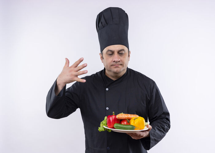 蔬菜男厨师身穿黑色制服 头戴厨师帽 手持盛有新鲜蔬菜的盘子 吸入新鲜蔬菜的香气 站在白色背景上香气戴着拿着
