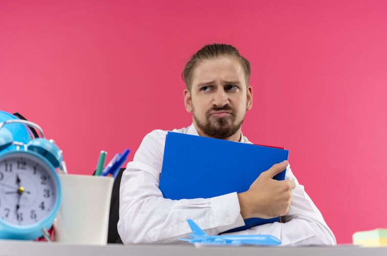 衬衫穿着白衬衫的帅哥商人拿着文件夹 带着悲伤的表情坐在粉色背景下的办公桌旁粉色桌子白色