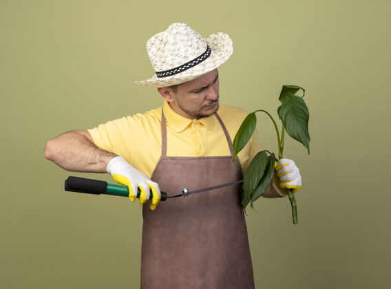 削减年轻的园丁 戴着工作手套 穿着连体衣 戴着帽子 手里拿着植物 用树篱剪剪着年轻人植物工作