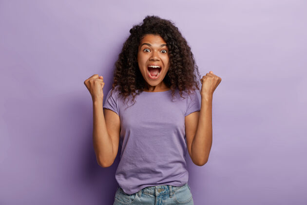 高兴情绪化的黑皮肤女人 非洲头发 举起紧握的拳头 兴奋地大叫 喜悦甜蜜的成功 感受胜利的味道 为最喜爱的球队呐喊 穿着紫色t恤和牛仔裤微笑紫罗兰年轻