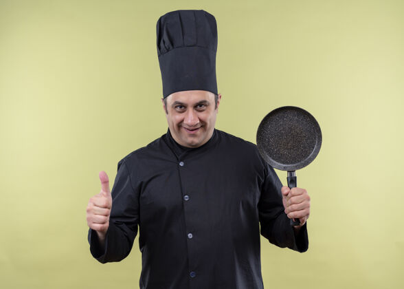 微笑男厨师身穿黑色制服 头戴厨师帽 手持平底锅 竖起大拇指 微笑着站在绿色背景上帽子站着男性