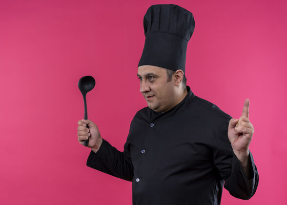 粉色男厨师身穿黑色制服 头戴厨师帽 手拿勺子 一边看一边露出食指 严肃的脸站在粉色背景上勺子拿着严肃
