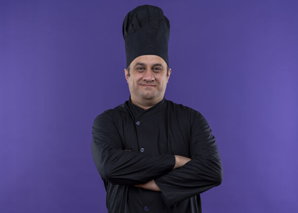 信心男厨师身穿黑色制服 头戴厨师帽 脸上带着自信的微笑 双手交叉放在胸前 站在紫色背景下看着镜头胸部烹饪微笑