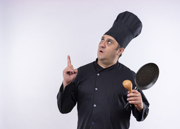惊喜男厨师身穿黑色制服 头戴厨师帽 手拿平底锅和木勺 食指朝上 惊讶地站在白色背景上帽子手指烹饪