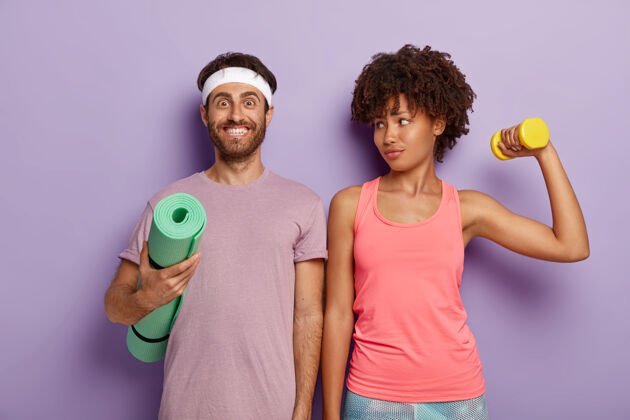 精力充沛笑容可掬的男人拿着健身垫摆姿势 穿着紫色t恤和头带 可爱的运动型女人看着丈夫 用体重训练二头肌 在室内肩并肩站着有氧运动和人姿势训练哑铃