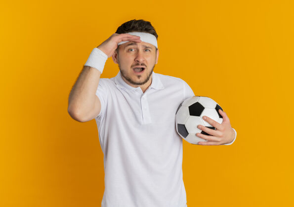足球身穿白衬衫 头戴足球带的年轻健身男子站在橙色背景下困惑地看着摄像机橙色白色头带