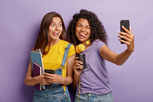 摄影师快乐的女人在一个小组里学习 在大学休息的时候玩得开心 在智能手机上自拍 展示舌头 拿着咖啡纸杯 拿着笔记本 一起对着紫色的墙摆姿势莫比尔娱乐饮料