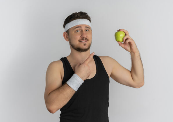 苹果戴着头巾的年轻健身男子拿着绿色的苹果 手指指着苹果 站在白色的背景上 开心地笑着手指白色头带