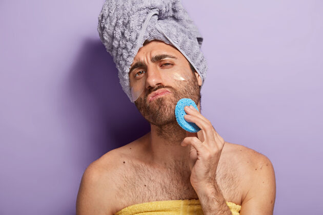 裸体放松严肃的男人用胡茬在淋浴后擦拭脸上的皮肤 拿着化妆海绵 用软毛巾包着治疗湿巾赤膊