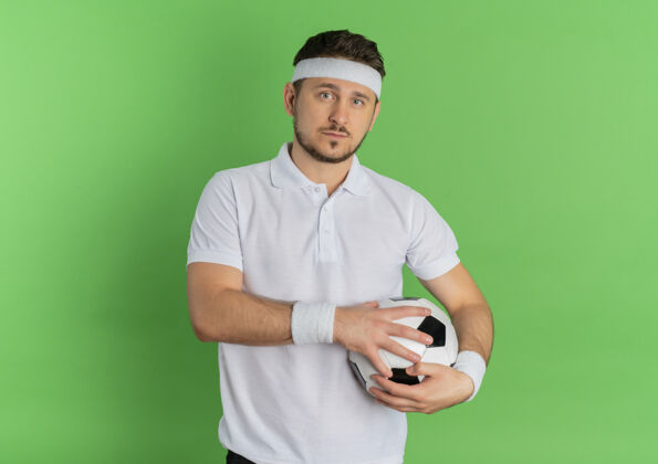 白色身穿白衬衫 头箍 手持足球的年轻健身男子站在绿色背景下 自信地看着摄像机看健身足球