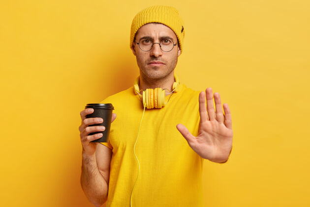 愤怒严重不满的家伙做了停止手势 拒绝做某事 说不 拿着外卖咖啡杯 戴着眼镜 穿着黄色休闲服男人耳机危险