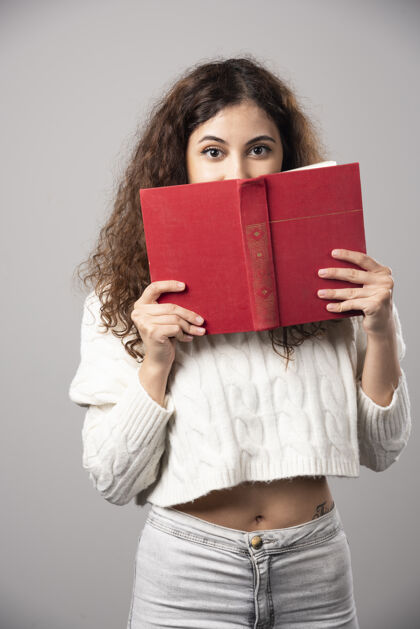 年轻一个年轻的女人在灰色的墙上用一本红色的书遮住她的脸高质量的照片女性肖像微笑