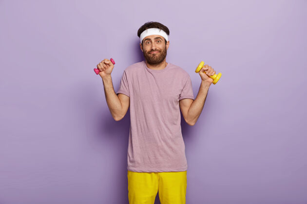紫色欧洲年轻人锻炼肌肉 用哑铃举起手臂 在室内锻炼 有运动的身体 穿着紫色t恤和黄色短裤 站在室内 有搞笑的样子 深色的鬃毛人胡茬男子