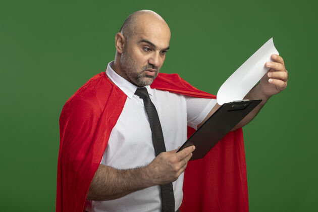 人穿红斗篷的超级英雄商人展示着空白页的剪贴板 站在绿色的墙壁上好奇地看着它商人红空白