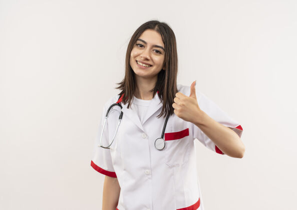 年轻人年轻的女医生穿着白大褂 脖子上戴着听诊器 面带微笑地站在白墙上竖起大拇指 望着前方周围女人听诊器