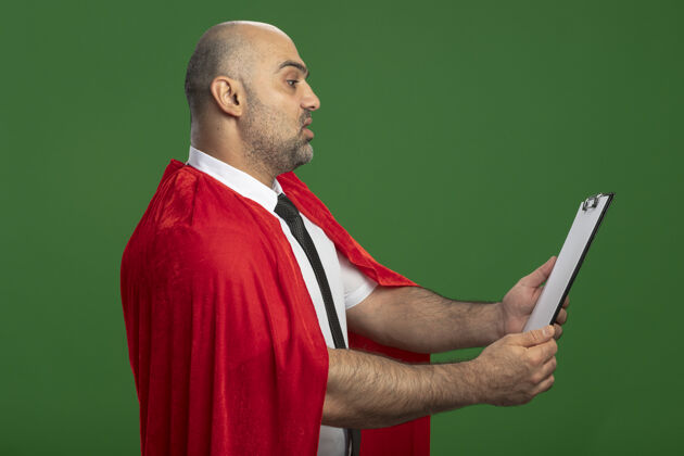 剪贴板穿红斗篷的超级英雄商人展示着空白页的剪贴板 严肃的脸站在绿色的墙上看着它显示超级超级英雄